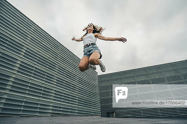 Carefree young woman jumping with arms outstretched against sky