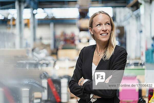 Lächelnde nachdenkliche Geschäftsfrau mit verschränkten Armen in einer Fabrik stehend