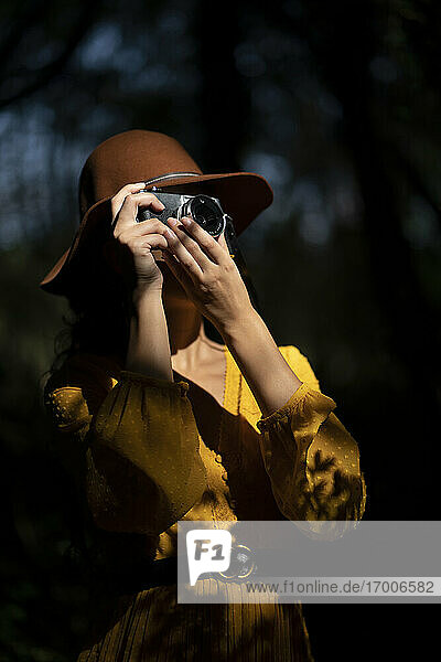 Junge Frau mit braunem Hut und gelbem Kleid  die ein Foto mit einer analogen Kamera im Wald macht