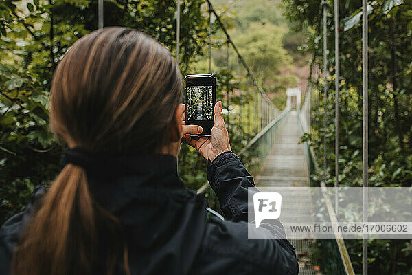Frau fotografiert mit Mobiltelefon auf Hängebrücke im Wald