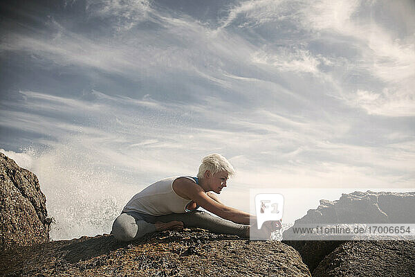 Blonde flexible Frau übt Yoga auf einer Felsformation am Strand gegen den Himmel