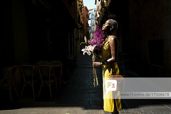 Woman with eyes closed holding bunch of flowers standing in city on sunny day