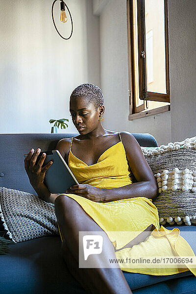 Frau im gelben Kleid benutzt digitales Tablet  während sie zu Hause auf dem Sofa sitzt