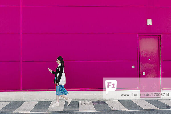 Young woman walking along pink wall  using smartphone