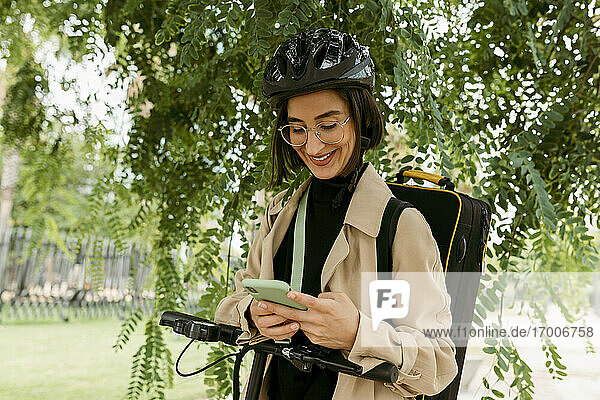 Lächelnde Frau mit Instrumentenkoffer und Elektroroller  die ein Mobiltelefon benutzt  während sie unter einem Baum im Park steht