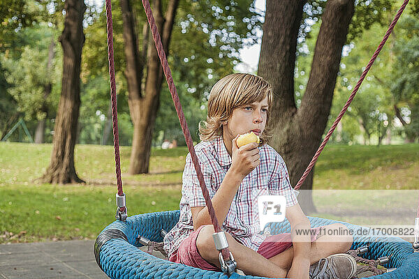 Nachdenklicher kleiner Junge isst Apfel  während er auf einer Seilschaukel im öffentlichen Park sitzt