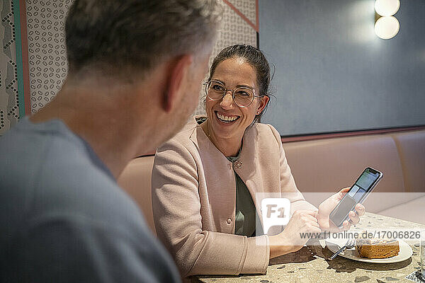 Businesswoman smiling while showing mobile phone to colleague while sitting at modern cafe