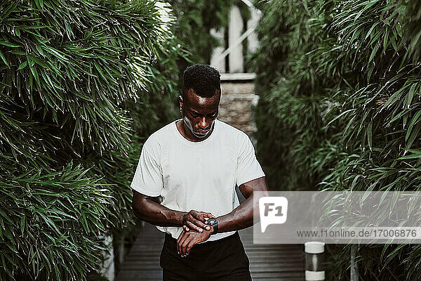 Männlicher Sportler überprüft seine Smartwatch  während er inmitten einer Pflanze steht