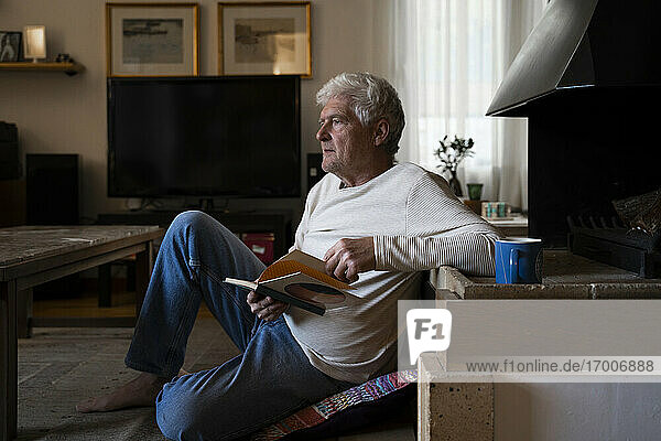 Nachdenklicher älterer Mann hält ein Buch  während er zu Hause auf dem Boden sitzt