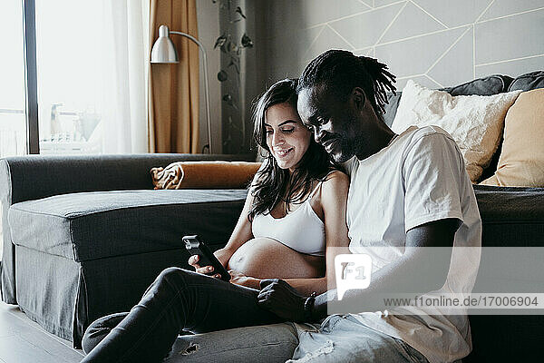 Junger Mann mit schwangerer Frau  die ein Smartphone benutzt  während sie zu Hause im Wohnzimmer sitzen