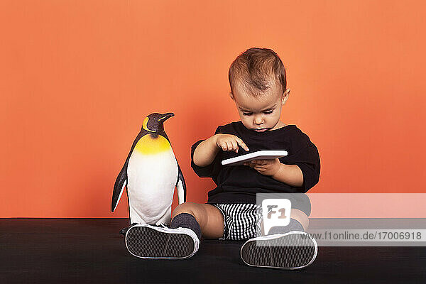 Baby-Mädchen  das ein Smartphone benutzt  während es neben einem Spielzeug vor einem orangefarbenen Hintergrund sitzt