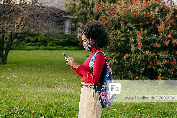 Studentin mit Kopfhörern und Gesichtsmaske  die im Freien steht und ihr Smartphone benutzt