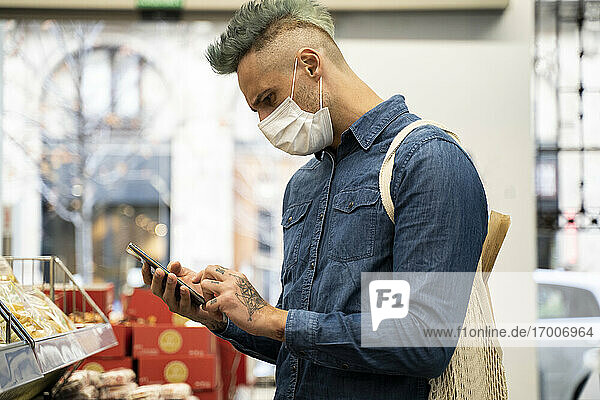 Stilvoller Mann mit Gesichtsmaske  der im Supermarkt ein Smartphone benutzt