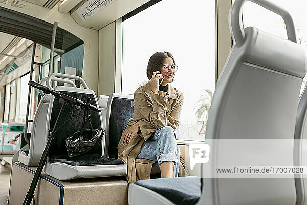 Smiling woman talking on mobile phone while sitting with electric push scooter in tram