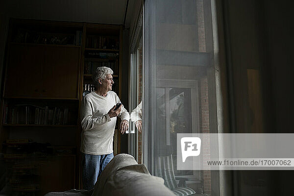 Älterer Mann mit Mobiltelefon  der wegschaut  während er zu Hause am Fenster steht