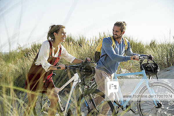 Smiling girlfriend and boyfriend walking with bicycles at beach