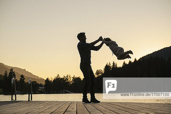 Silhouettes of father and little daughter playing at end of lakeshore jetty at dusk