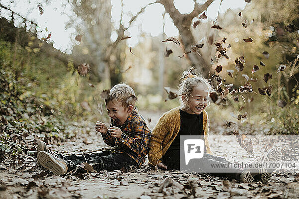 Fröhliches Mädchen und Junge spielen mit trockenen  gefallenen Blättern  während sie auf einem Fußweg im Wald sitzen