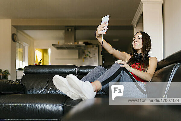 Smiling teenage girl taking selfie through mobile phone while sitting on sofa at home