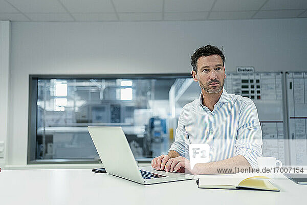 Älterer Geschäftsmann mit Laptop  der im Sitzungssaal eines Industriebüros nachdenkt