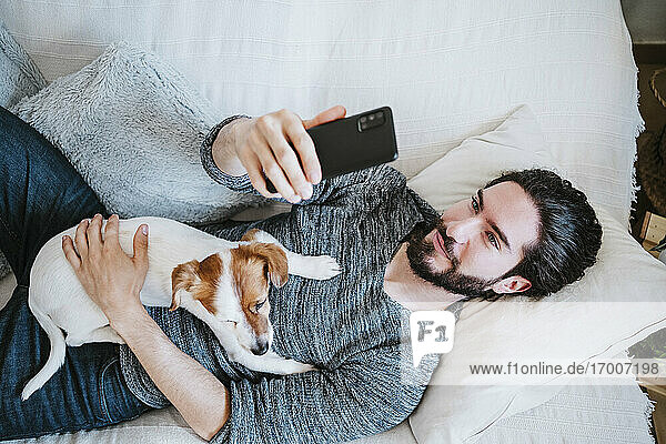 Young man taking selfie through mobile phone while lying on sofa with dog at home