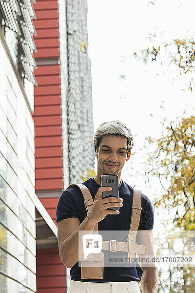 Lächelnder Mann mit Hand in der Tasche  der sein Smartphone in der Stadt benutzt