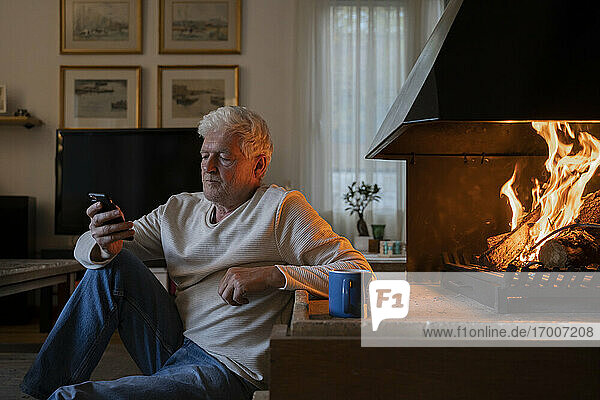 Ein älterer Mann benutzt ein Mobiltelefon  während er zu Hause am Kamin sitzt