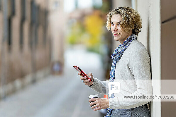 Lächelnder Mann mit Einwegbecher  der sein Handy benutzt und sich an die Wand lehnt