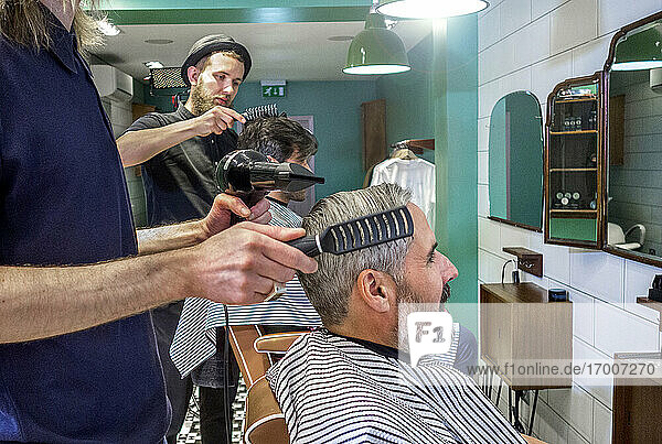 Male hairdresser using dryer while combing customer's hair at barber shop