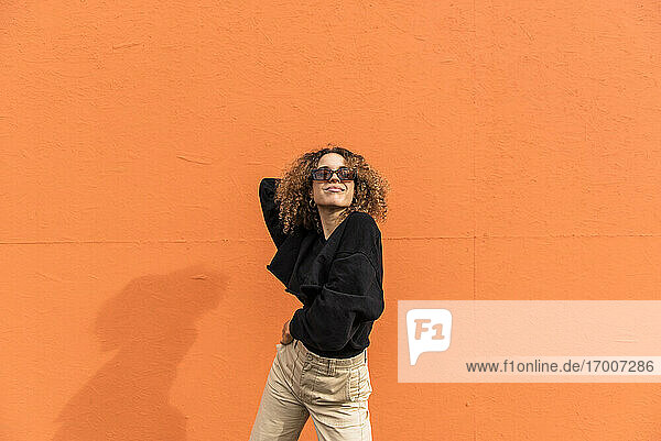 Junge Frau mit Sonnenbrille tanzt  während sie gegen eine orangefarbene Wand steht