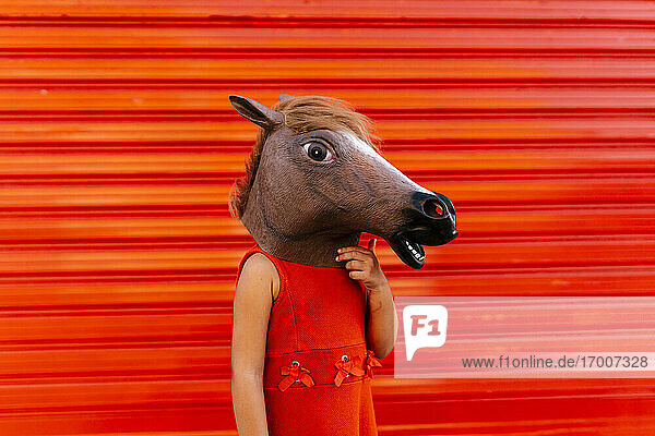 Kleines Mädchen mit einem Pferdekopf und einem roten Kleid vor einem roten Rollladen