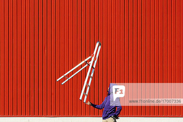 Young man balancing a ladder in front of a red wall