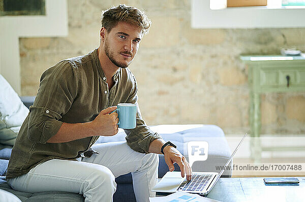 Männlicher Freiberufler hält Kaffeetasse  während er zu Hause am Laptop sitzt