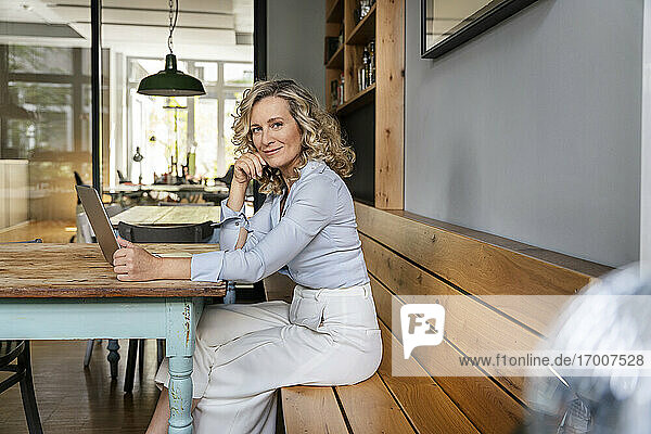 Lächelnde Geschäftsfrau mit Hand am Kinn  die mit einem Laptop am Tisch in einer Büro-Cafeteria sitzt