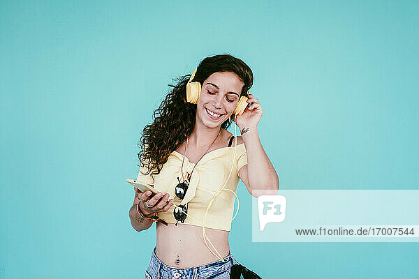 Smiling beautiful woman with eyes closed listening music through headphones against blue wall