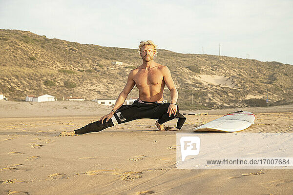 Shirtless man stretching by surfboard on sand at beach