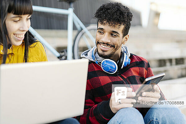Lächelnde Freunde  die im Freien sitzend einen Laptop und ein digitales Tablet benutzen