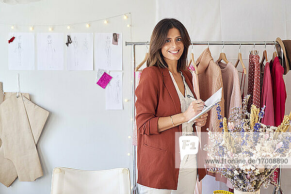 Smiling beautiful fashion designer writing in diary while standing against clothes rack at atelier