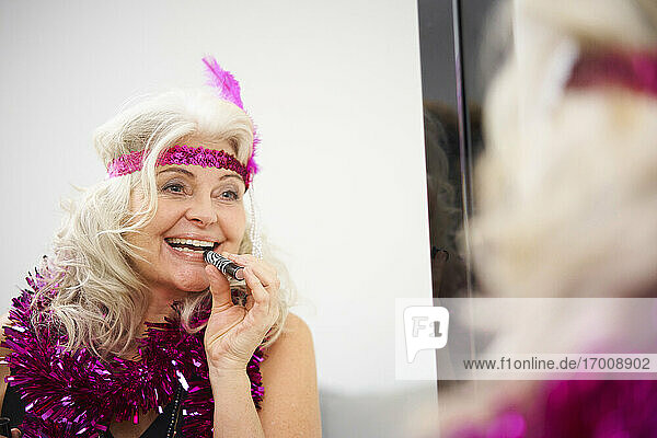 Happy senior woman with garland around neck applying lipstick in front of mirror at home