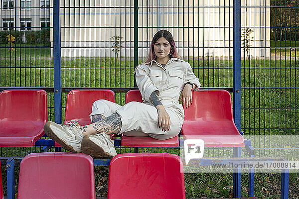 Junge Frau posiert  während sie auf einem Sitz gegen den Zaun eines Sportplatzes sitzt