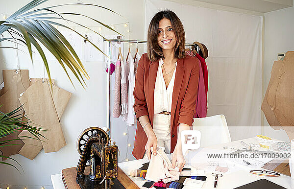 Lächelnde Designerin  die ein Stoffmuster am Schreibtisch vor einem Kleiderständer in einem Atelier hält