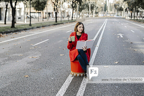 Junge Frau liest ein Buch und hält eine Kaffeetasse auf einem Sitz an der Straße
