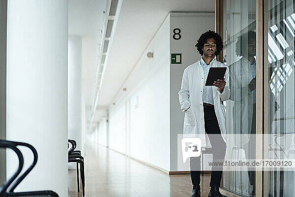 Selbstbewusster junger Arzt  der ein digitales Tablet benutzt  während er sich an eine Glaswand im Krankenhauskorridor lehnt