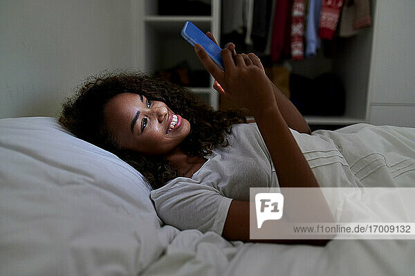 Happy young woman using smart phone while lying on bed in bedroom at home