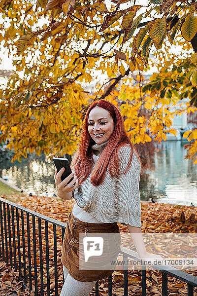 Schöne Frau  die ihr Smartphone benutzt  während sie im Herbst an einem Geländer im Park steht
