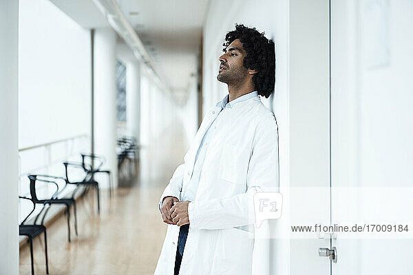 Thoughtful young male healthcare worker leaning on white wall at hospital corridor