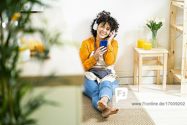 Junge Frau mit Kopfhörern  die ein Mobiltelefon benutzt  während sie zu Hause auf dem Boden sitzt