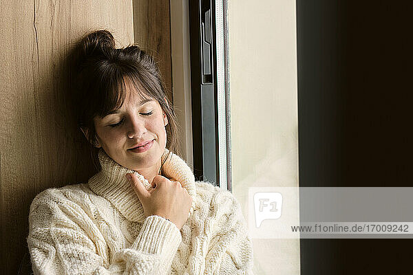 Thoughtful woman with eyes closed sitting by window at home during winter