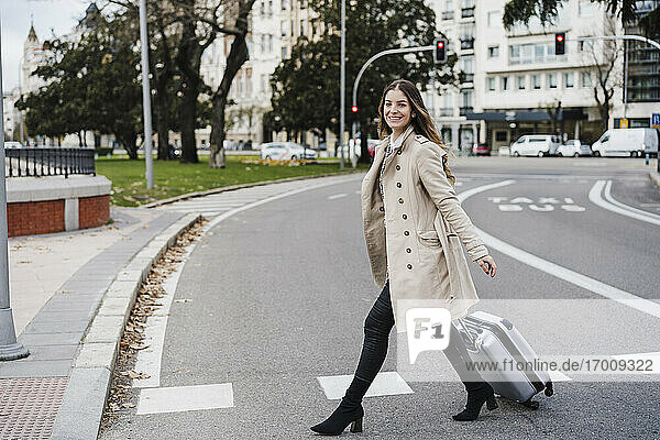 Fröhliche Frau mit Gepäck auf Rädern beim Überqueren der Straße in der Stadt