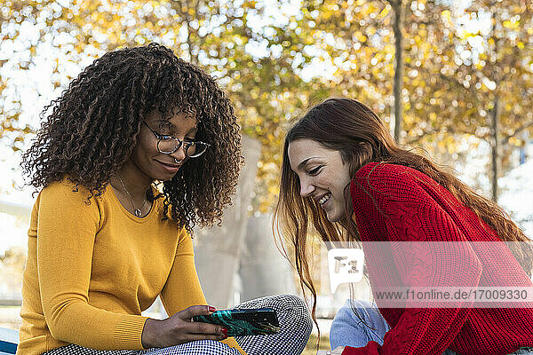 Lächelnde junge Frauen  die in einem öffentlichen Park auf ihr Mobiltelefon schauen